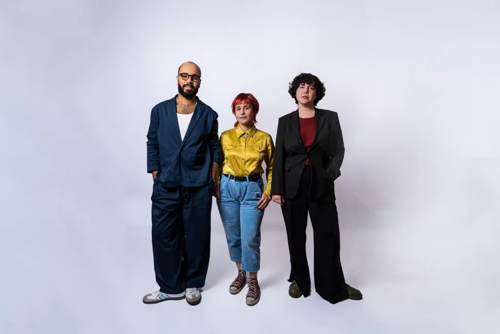 Três pessoas estão posando lado a lado diante de um fundo branco. À esquerda, uma pessoa de barba, óculos e cabeça raspada veste conjunto azul-escuro com camiseta branca e tênis claros. Ao centro, uma pessoa de cabelo curto avermelhado usa camisa amarela brilhante, calça jeans dobrada e tênis marrons. À direita, uma pessoa de cabelo curto e cacheado veste terno preto com camiseta vinho e sapatos verde-oliva. Todos olham para a câmera com expressões neutras.