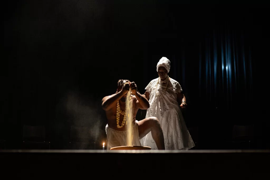 Cena teatral em um palco escuro, iluminado por uma luz suave. Em primeiro plano, uma pessoa ajoelhada despeja um punhado de grãos ou farinha em uma bacia, criando um efeito de poeira no ar. Ela veste um tecido claro na cintura e colares grandes. Atrás dela, outra pessoa, vestida com roupas tradicionais brancas e um torço na cabeça, observa a ação com postura serena. A atmosfera é ritualística e intensa, com um clima de espiritualidade e concentração.